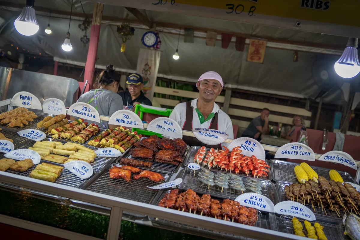 Vendor behind a food counter with skewers and menu signs at Bang Niang Market