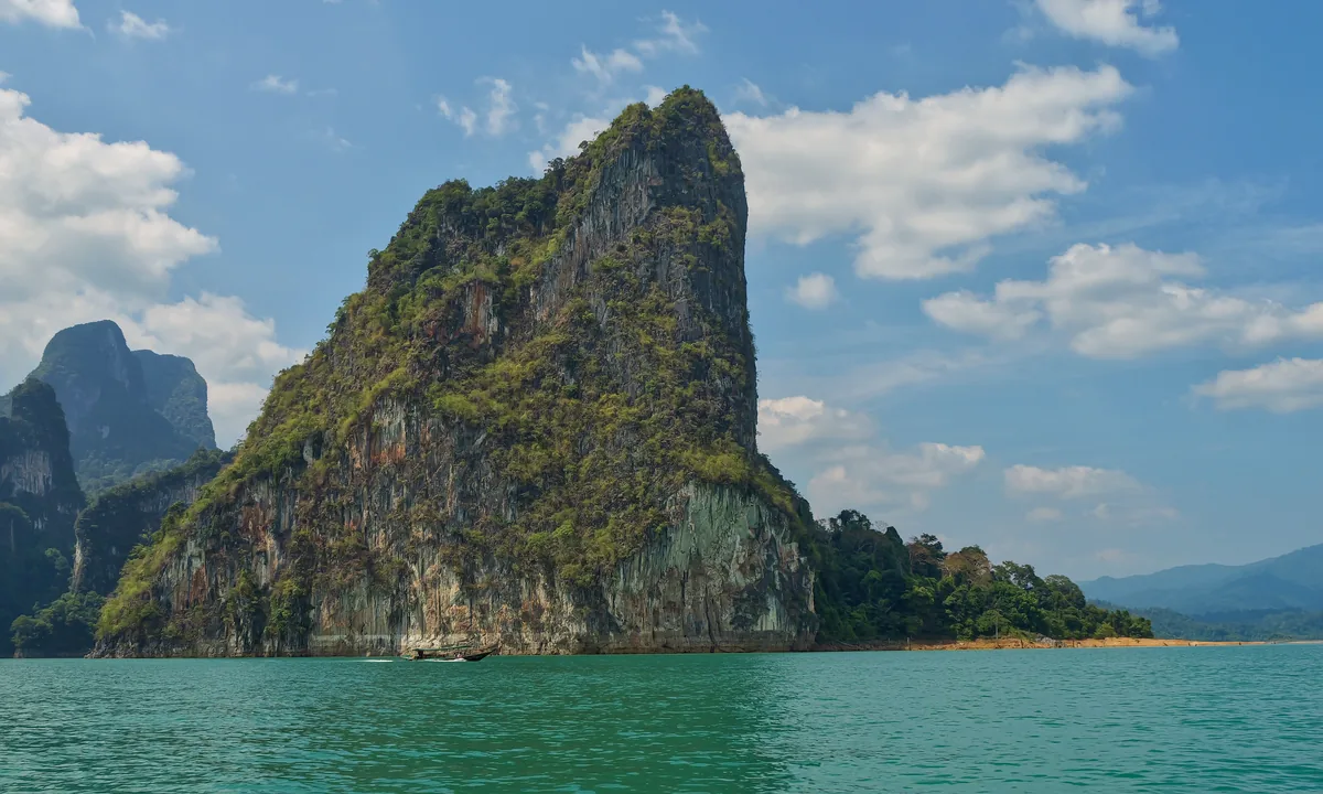 Steep forested cliffs rising above green water at Cheow Lan Lake, with a small boat below