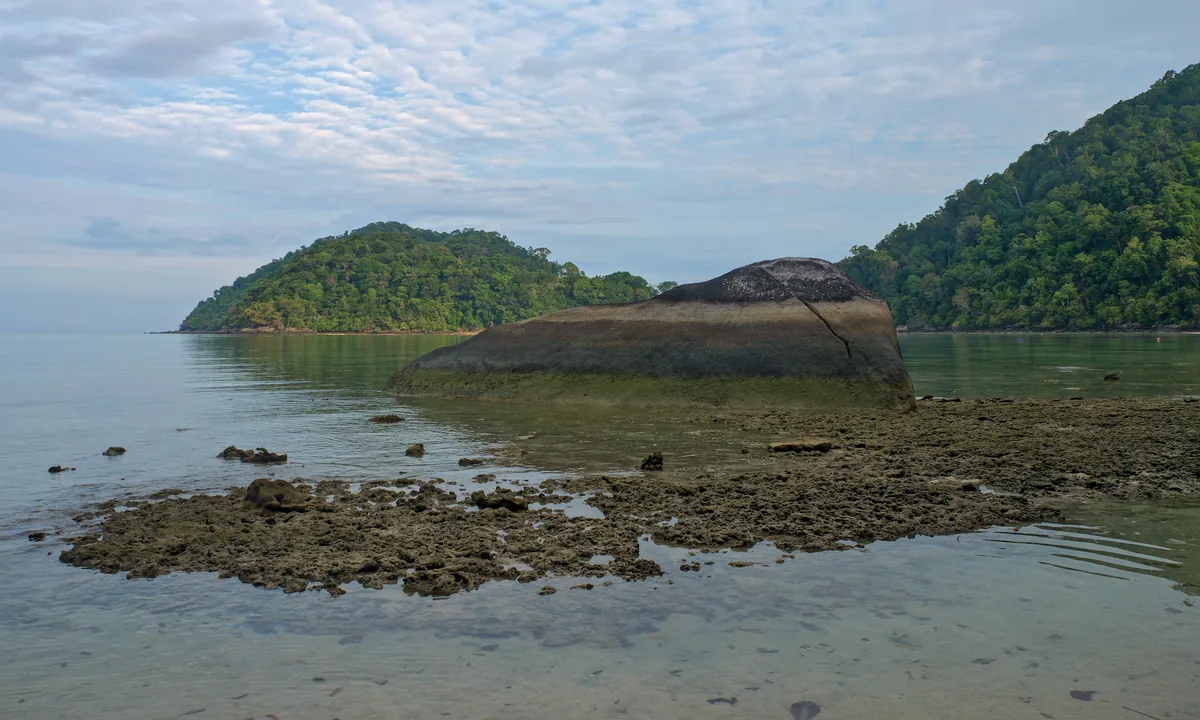Large boulder in shallow water with forested shores behind it on North Surin Island